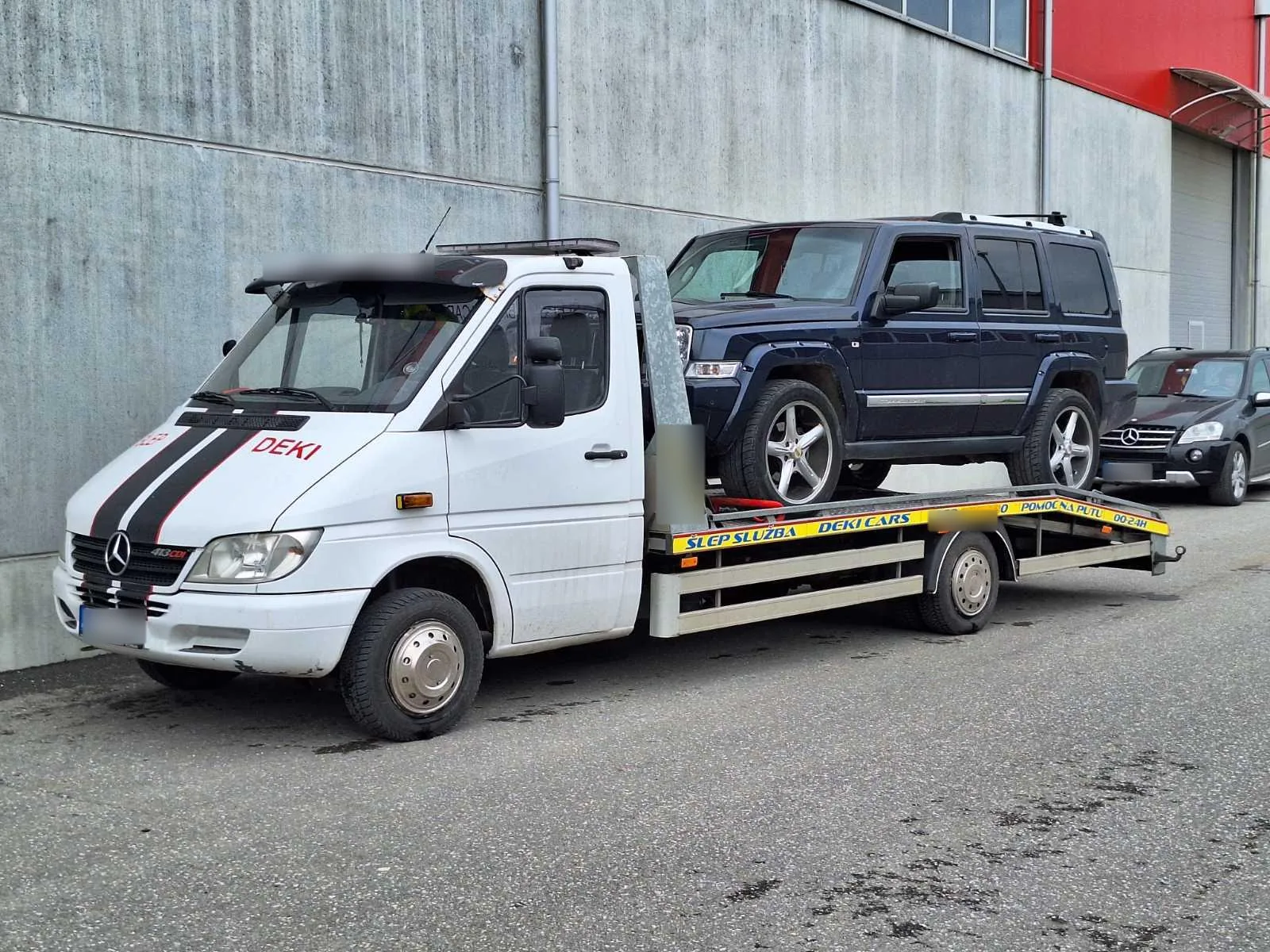 Šlep vozilo Deki Team sa tamnim terenskim automobilom ispred industrijske hale.